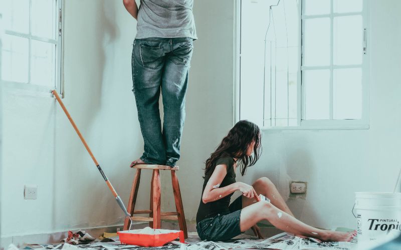 Woman sitting on the floor next to a man that's standing on a stool while painting their apartment