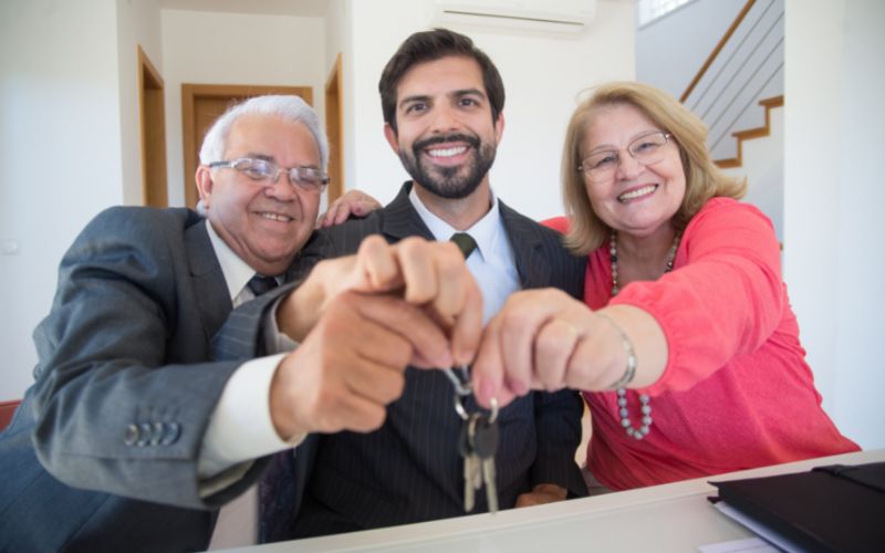 An elderly couple on each side of a real estate agent holding home keys in front of the camera.